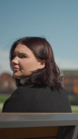 close up de la dama sentada en un columpio de madera al aire libre, su cabello se mueve ligeramente con la brisa, con una vista borrosa de un edificio y vegetación en el fondo en un día soleado brillante