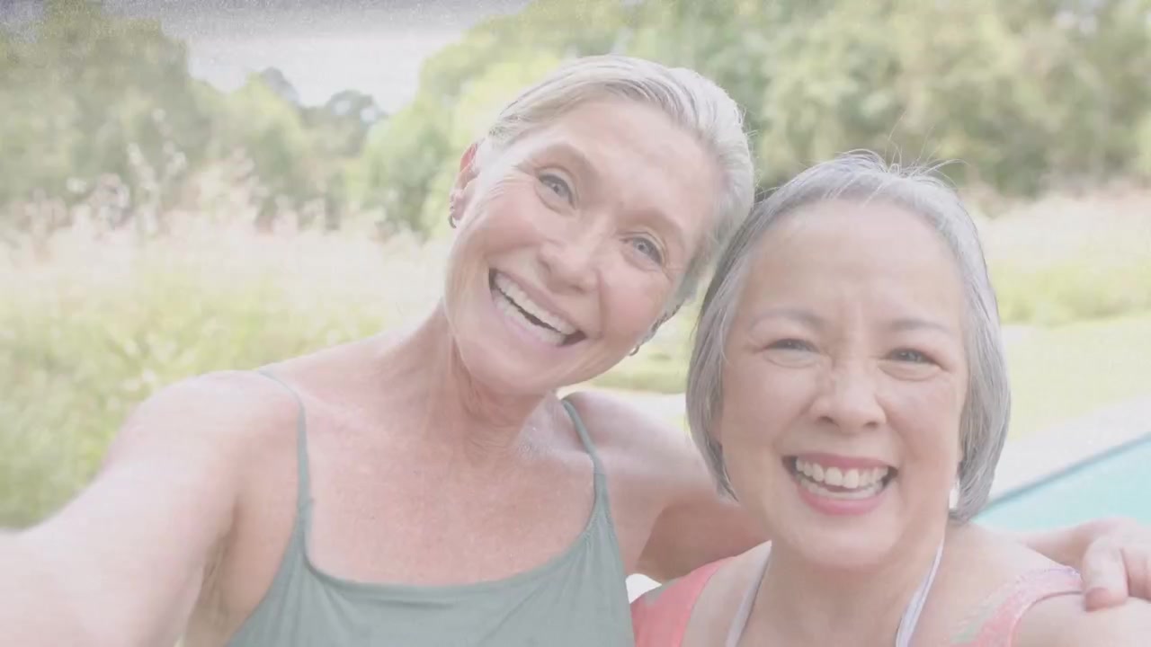 腕を組んで笑い合う2人の女性が、公園でスマートフォンを伸ばし、ソーシャルメディア用の自撮りを安定させている。粒状のオーバーレイがかかっている。