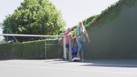 smiling senior caucasian couple, arriving to play tennis at sunny outdoor tennis court, slow motion