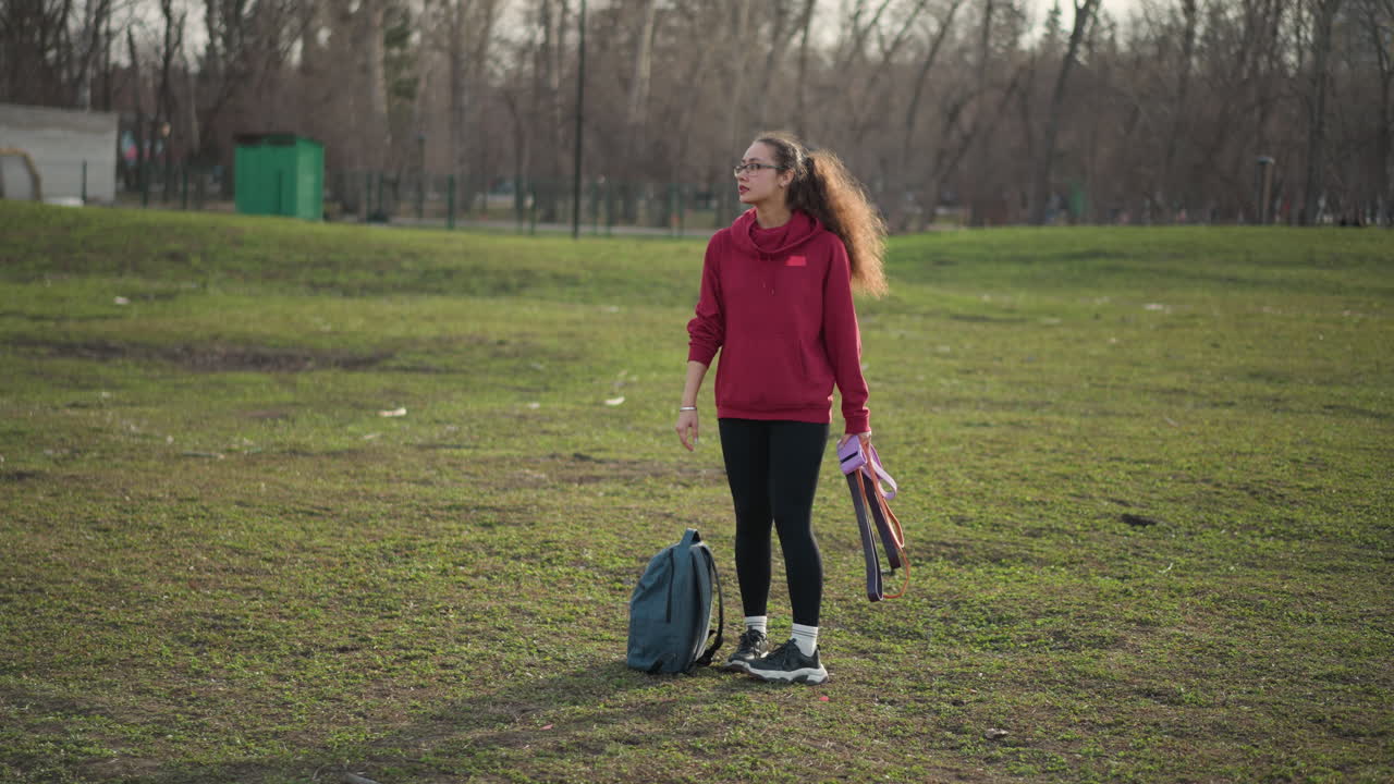 Woman Exercising With Bands Outside, Energetic Woman Engaged In Outdoor Strength Training Session, Athletic Woman Methodically Exercising Outside While Wearing Red Hoodie And Carrying Backpack