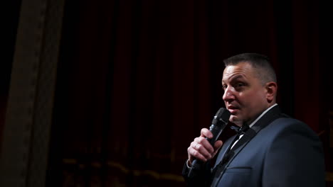 experienced host with a microphone. positive man in gray costume standing on theatrical stage and talking on camera. lockdown during pandemic.