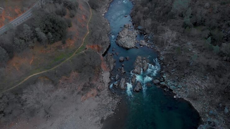 Drohnenflug hoch über dem blauen American River mit Bergblick auf Auburn, Kalifornien, bei Sonnenuntergang – umgeben von grünen Bäumen und Bergen.