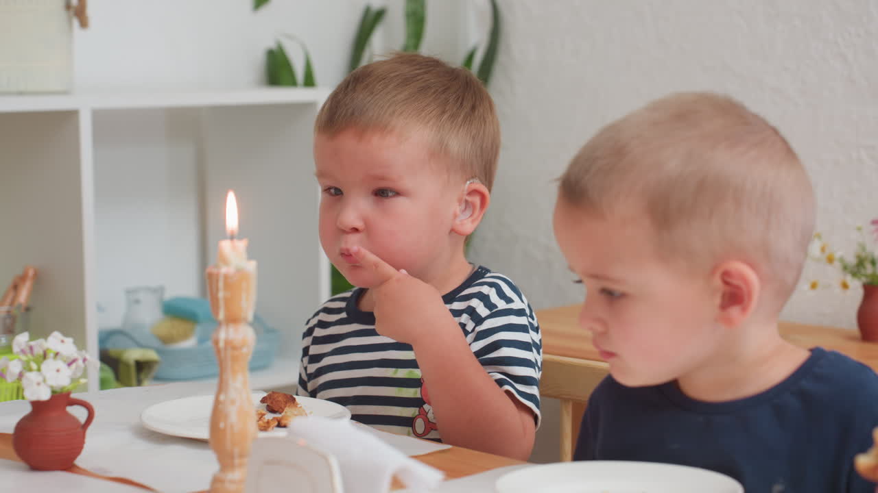 Two young boys sitting at wooden table during kindergarten meal, one wearing striped shirt with hearing aid, looking thoughtful while other eats quietly under soft daylight with candle