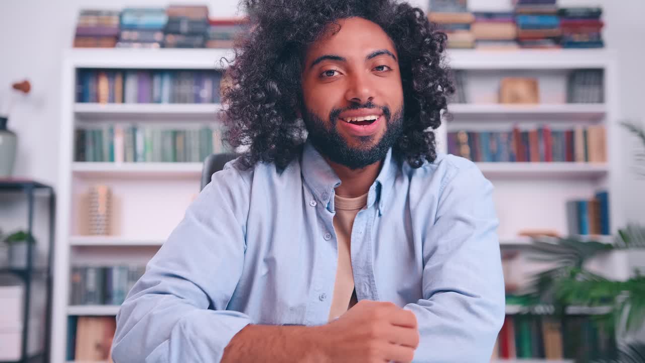 Bell'uomo arabo che sorride alla telecamera, seduto al tavolo in biblioteca o in uno studio.