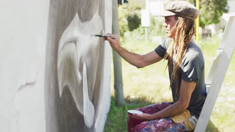 Video-of-caucasian-male-artist-with-dreadlocks-sitting-on-ladder-painting-whale-mural-on-wall