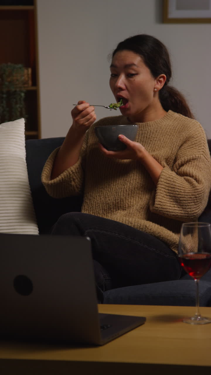 video vertical de una mujer en casa sentada en el sofá comiendo una comida saludable vegetariana o vegana de ensalada tabbouleh con un vaso de vino viendo el programa en la computadora portátil 1