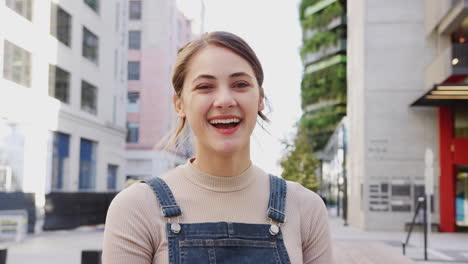 Trendy-young-Hispanic-woman-wearing-dungarees-sitting-in-the-street-laughing-to-camera,-close-up