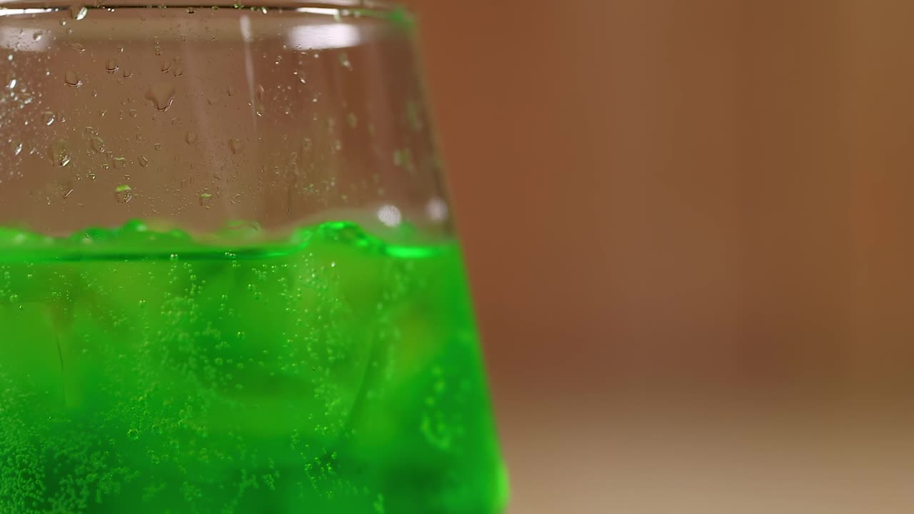 Close-up of a bright green beverage with ice cubes in a transparent glass against a blurred background.