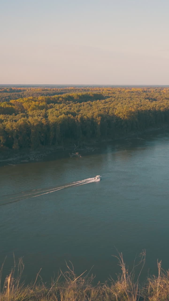 modern motorboat sails along wide calm river past dense forest against clear blue sky on nice autumn evening