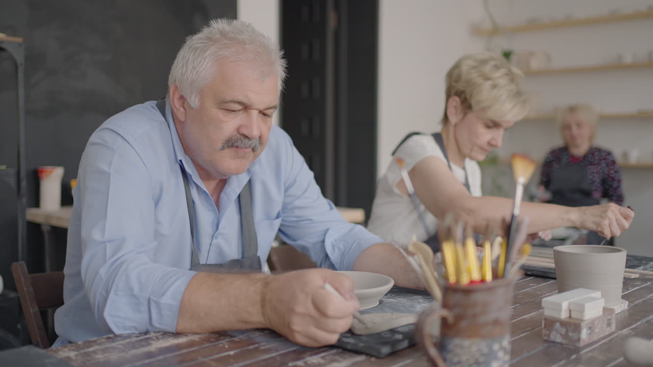A group of elderly people at a master class in pottery together sculpt and cut a drawing on cups of clay for the manufacture of ceramic dishes