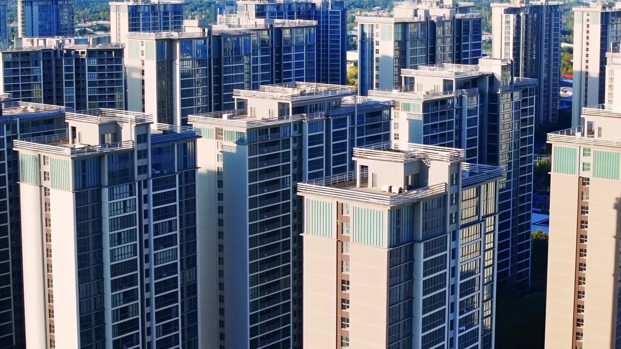 ガラス張りのバルコニーと明るいコンクリートのファサードを持つ、密集した現代的な高層マンション群を右側から見た真昼の空撮。強い日差し