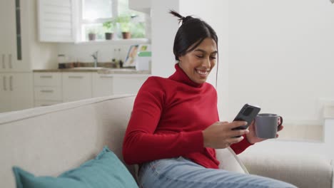 Video-of-happy-biracial-woman-in-hijab-sitting-on-sofa-and-using-smartphone