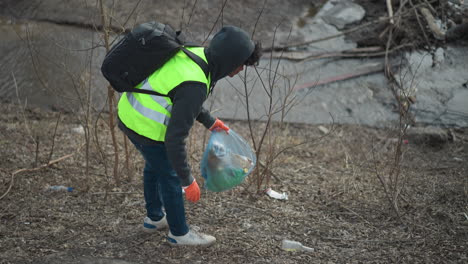 volunteer in reflective vest and gloves crouches to collect litter from ground near riverbank, placing trash into plastic bag during environmental cleanup to protect nature and promote community