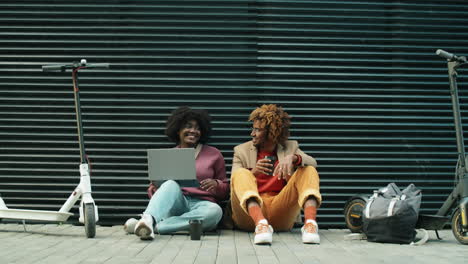 african american friends using laptop and talking while sitting on street