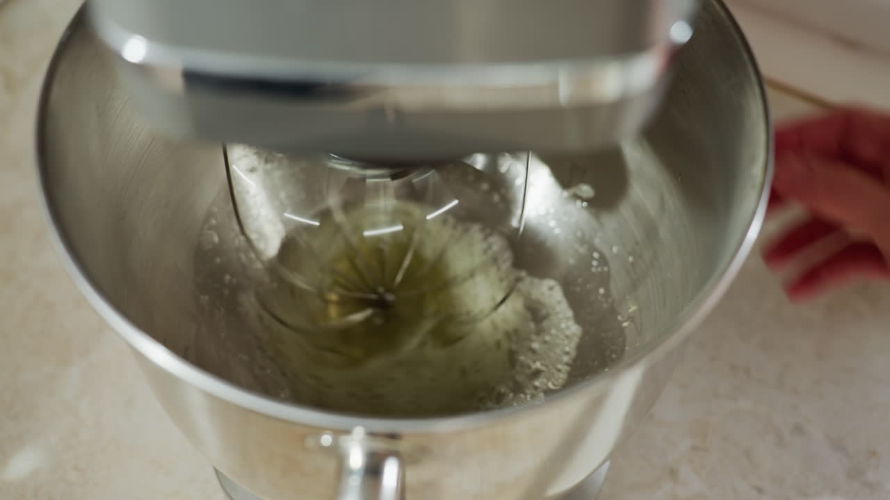 Close up of hand turning on digital electric mixer as stainless whisk begins spinning inside metal bowl with egg liquid, showcasing kitchen appliance in action during food preparation process