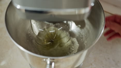 close up of hand turning on digital electric mixer as stainless whisk begins spinning inside metal bowl with egg liquid, showcasing kitchen appliance in action during food preparation process