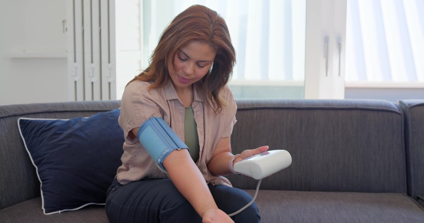 Video verticale: Monitor che si avvia, donna con un top beige che regola il bracciale, misurando la pressione sanguigna sul divano.