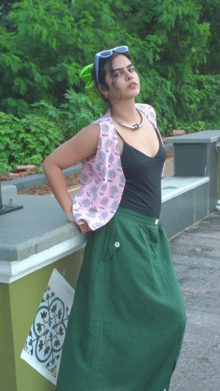 Full-length vertical portrait of a young woman in a green skirt, black top, and sunglasses posing playfully outdoors by a tiled bench, with lush green foliage in background, relaxed stylish look