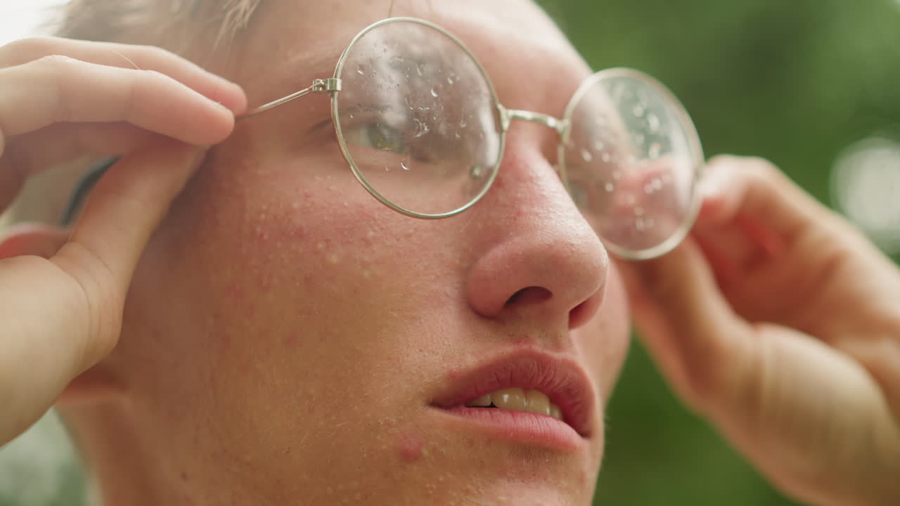 Ung hvid mand justerer våde briller, runde glas besprøjtet med regndråber, nærbillede i grøn have, vintage briller, reflekterende blik, studerende holder pause mellem timerne, blødt sollys
