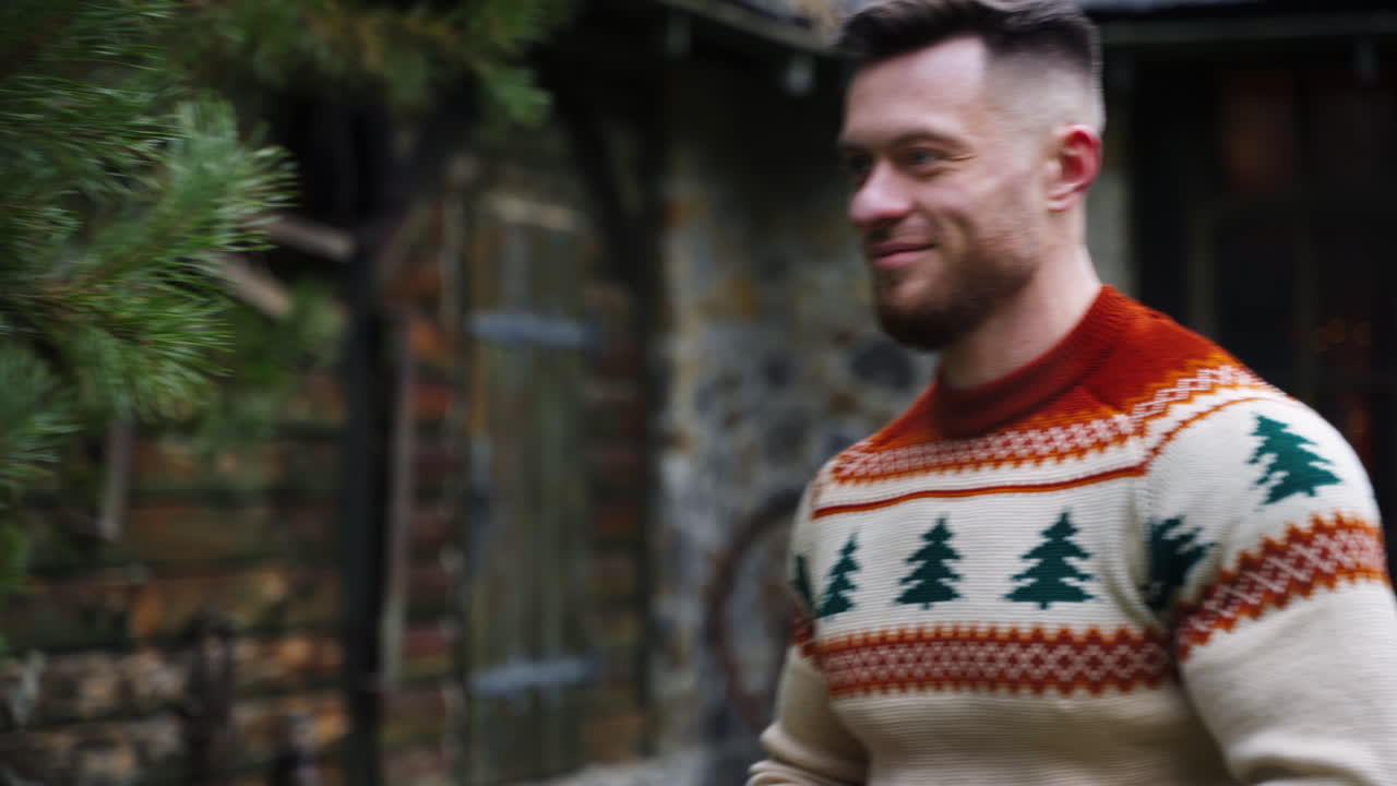Smiling young guy on cottage background. Handsome man in warm sweater walking to someone in terrace. Guy with a warm blanket in hands outdoors.