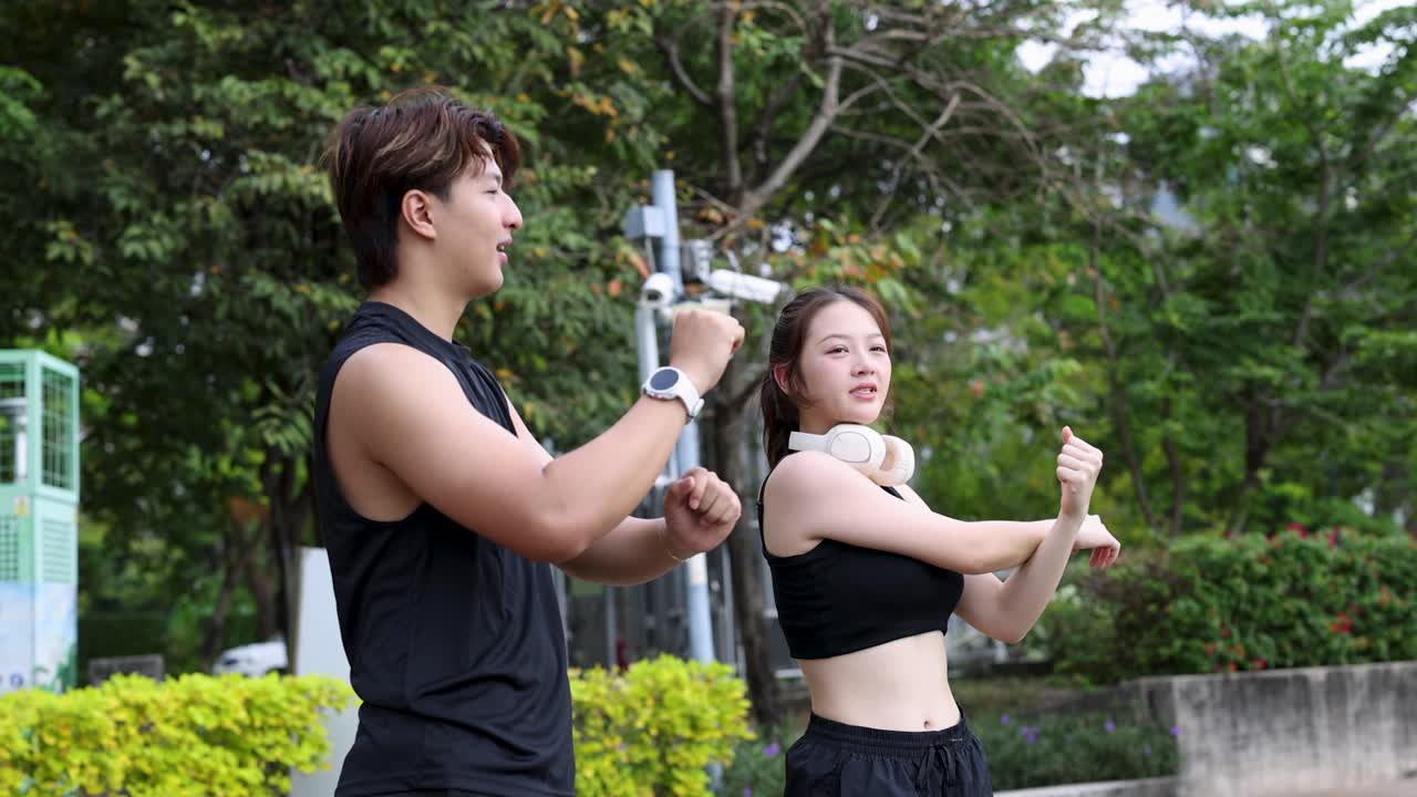 Một chàng trai và một cô gái mặc đồ thể thao màu đen đang thực hiện các động tác giãn cơ tay trong công viên
