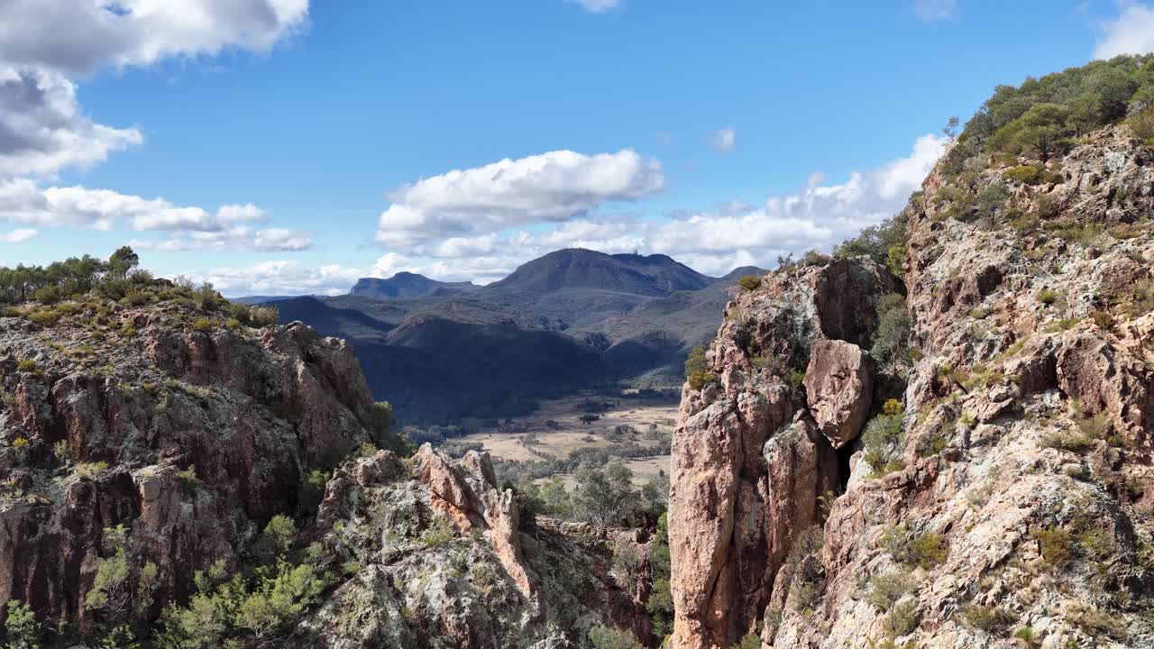 Daylight camera pan reveals rugged Split Rock cliffs, expansive forested valleys, and distant mountains under blue skies in Warrumbungle National Park, Australia