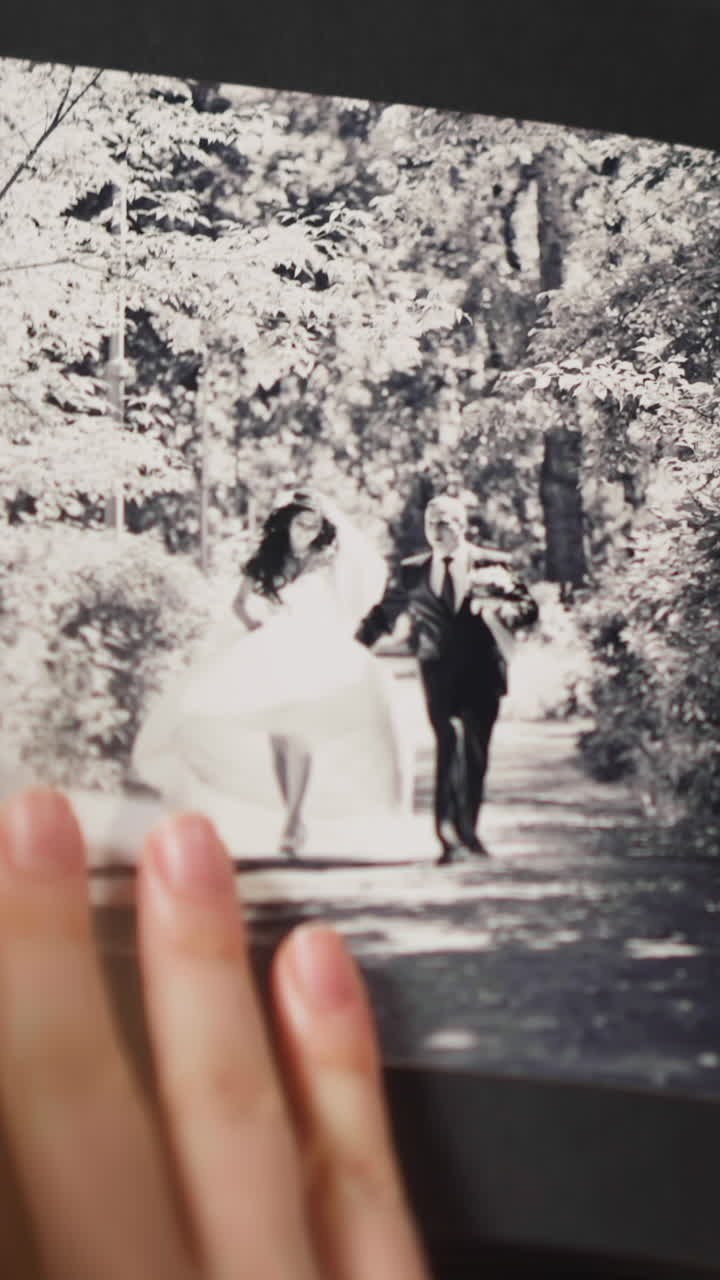 Woman looks at vintage black and white faded picture of grandparents wedding in large photobook at table in light room closeup slow motion