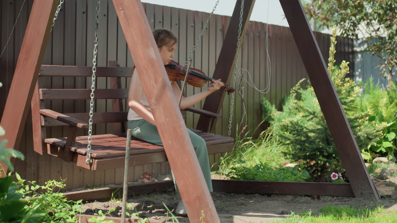 Ragazza con violino all'aperto, ragazza con archetto in un giardino soleggiato che suona il violino sull'altalena, ragazza con abbigliamento estivo casual che si esercita attentamente all'aperto in mezzo al verde lussureggiante e alle recinzioni