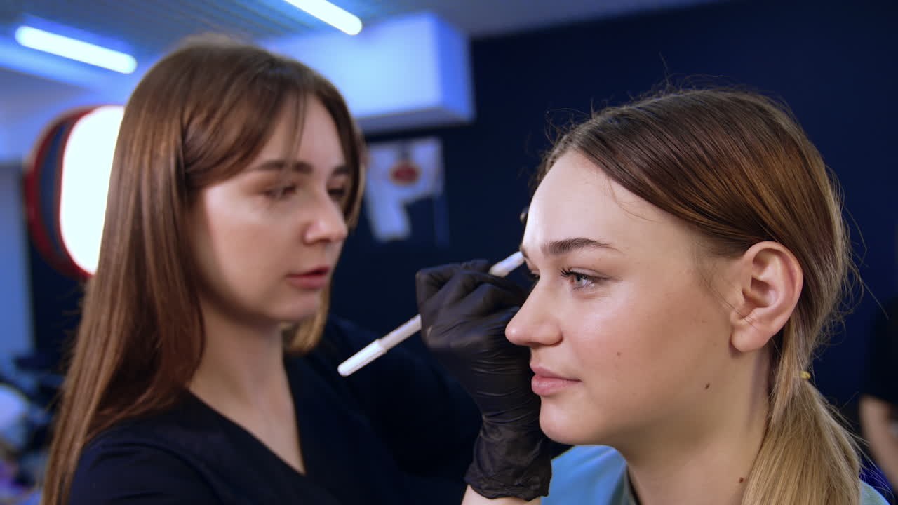 Young beautiful Caucasian girl is at salon. Brunette long-haired brow master drawing lines for future eyebrows. Close up.