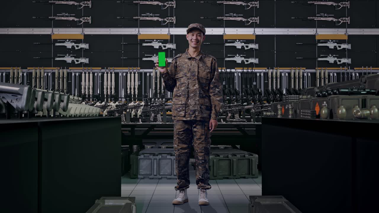 Soldato asiatico sorridente mostra uno smartphone con schermo verde alla telecamera in un arsenale militare