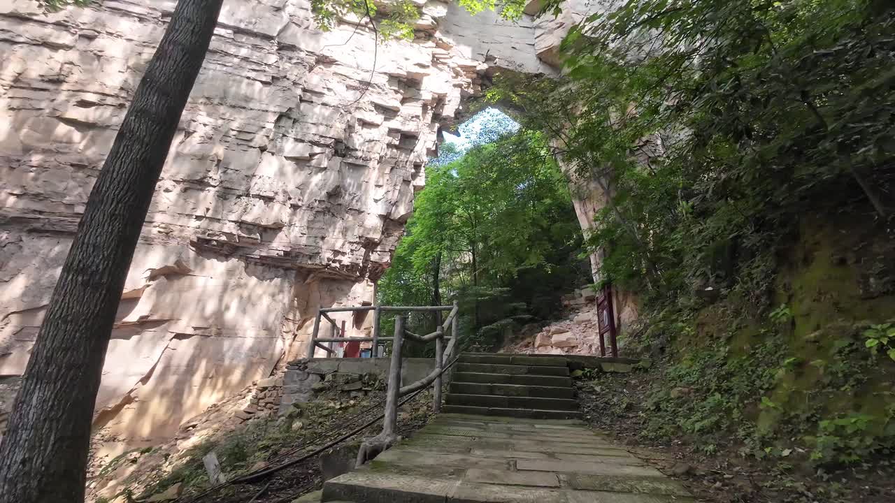 Textured cliff face and walkway in Zhangjiajie surrounded by lush vegetation, camera pans right