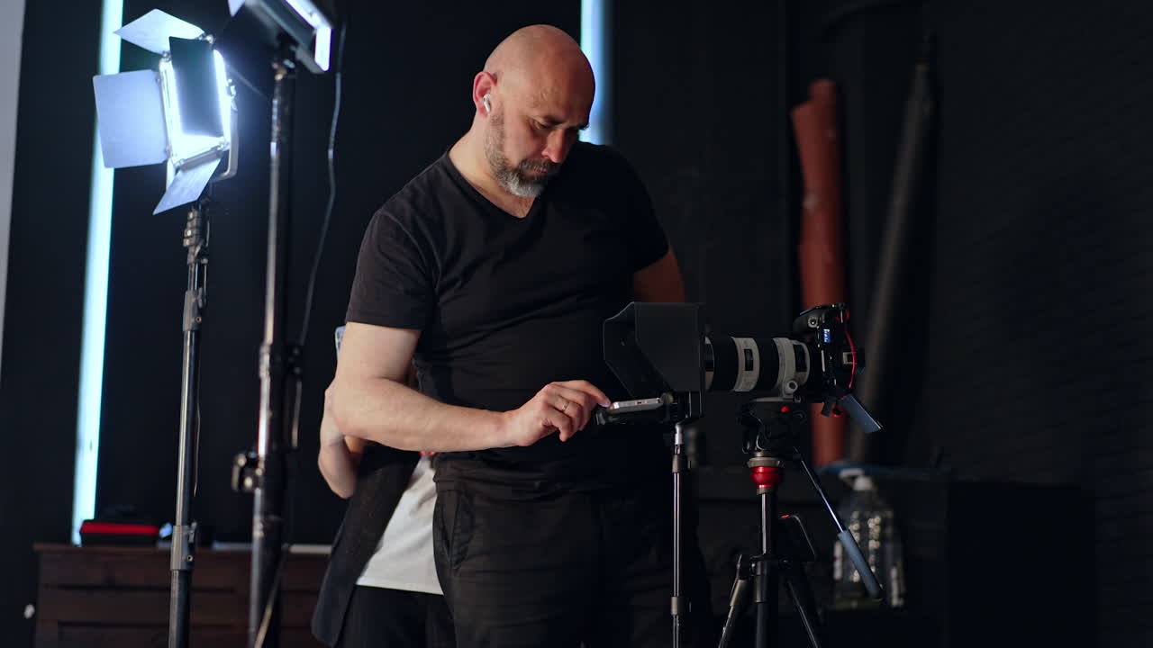 Bearded bald photographer stands at the camera on tripod. Cameraman sets the video prompter. Long-haired girl stands nearby focused on her phone.