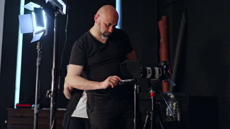 bearded bald photographer stands at the camera on tripod. cameraman sets the video prompter. long-haired girl stands nearby focused on her phone.