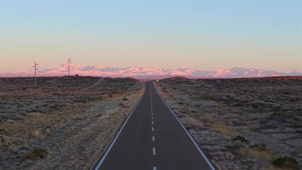 Vue de drone en suivi latéral gauche au-dessus de l'autoroute Route 144, traversant des champs arides et avec en toile de fond un lever de soleil éclairé de violet sur la chaîne de montagnes des Andes