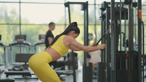 una joven hispana en un chándal amarillo realiza un ejercicio en un crossover tira de una cuerda desde arriba para entrenar la espalda y los hombros. una mujer morena entrena la espalda y los hombros en el gimnasio