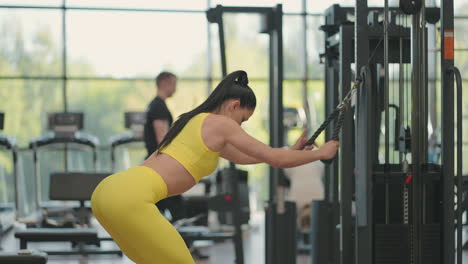 a young hispanic woman in a yellow tracksuit performs an exercise in a crossover pulls a rope from above to train the back and shoulders. brunette woman trains back and shoulders in the gym