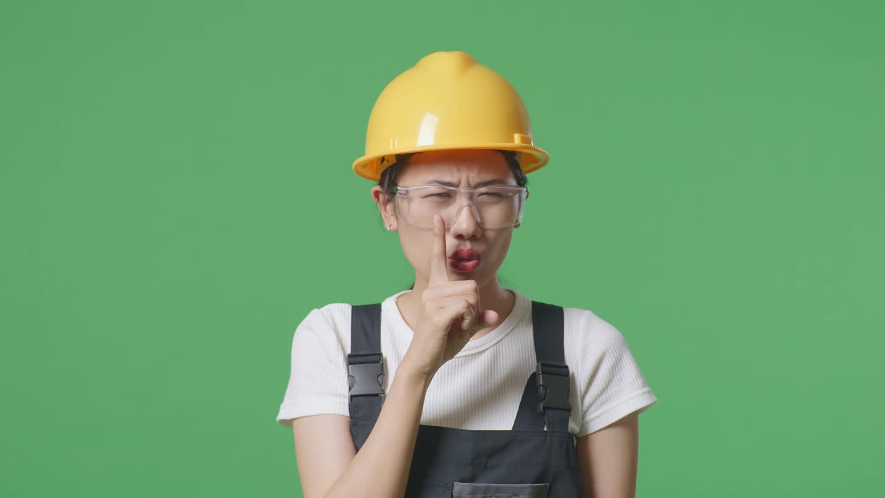 안경과 안전 헬을 착용한 아시아 여성 노동자가 녹색 스크린 배경 스튜디오에 서서 카메라를 바라보고 shh 제스처를합니다.
