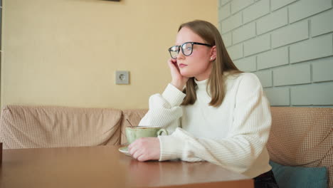 lonely investor in white sweater seated indoors stirring coffee slowly while resting hand under jaw in quiet contemplation, wall with artwork visible in soft background of peaceful cafe corner