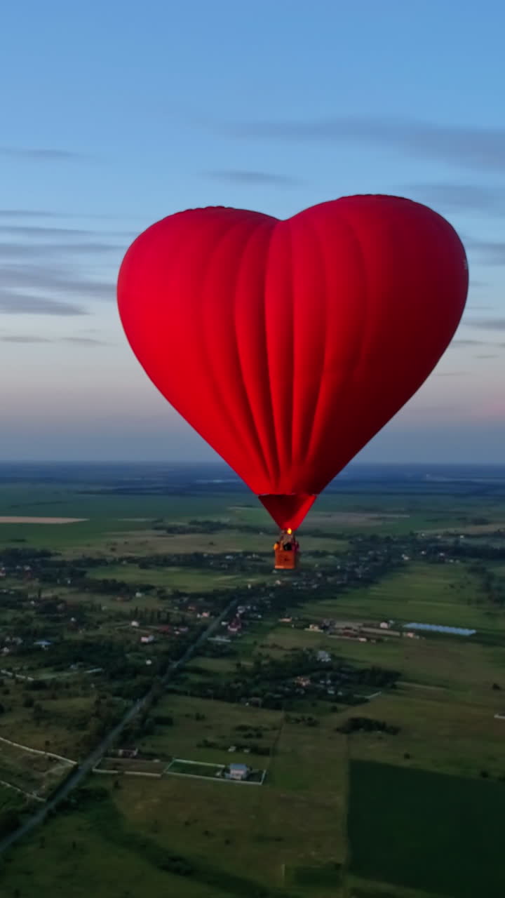 Red airship in the shape of a heart. Hot air balloon in the sky over a field in the countryside flying in the beautiful evening sky. Vertical video