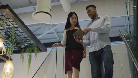 video of two diverse male and female colleagues talking and using tablet on stairs at office