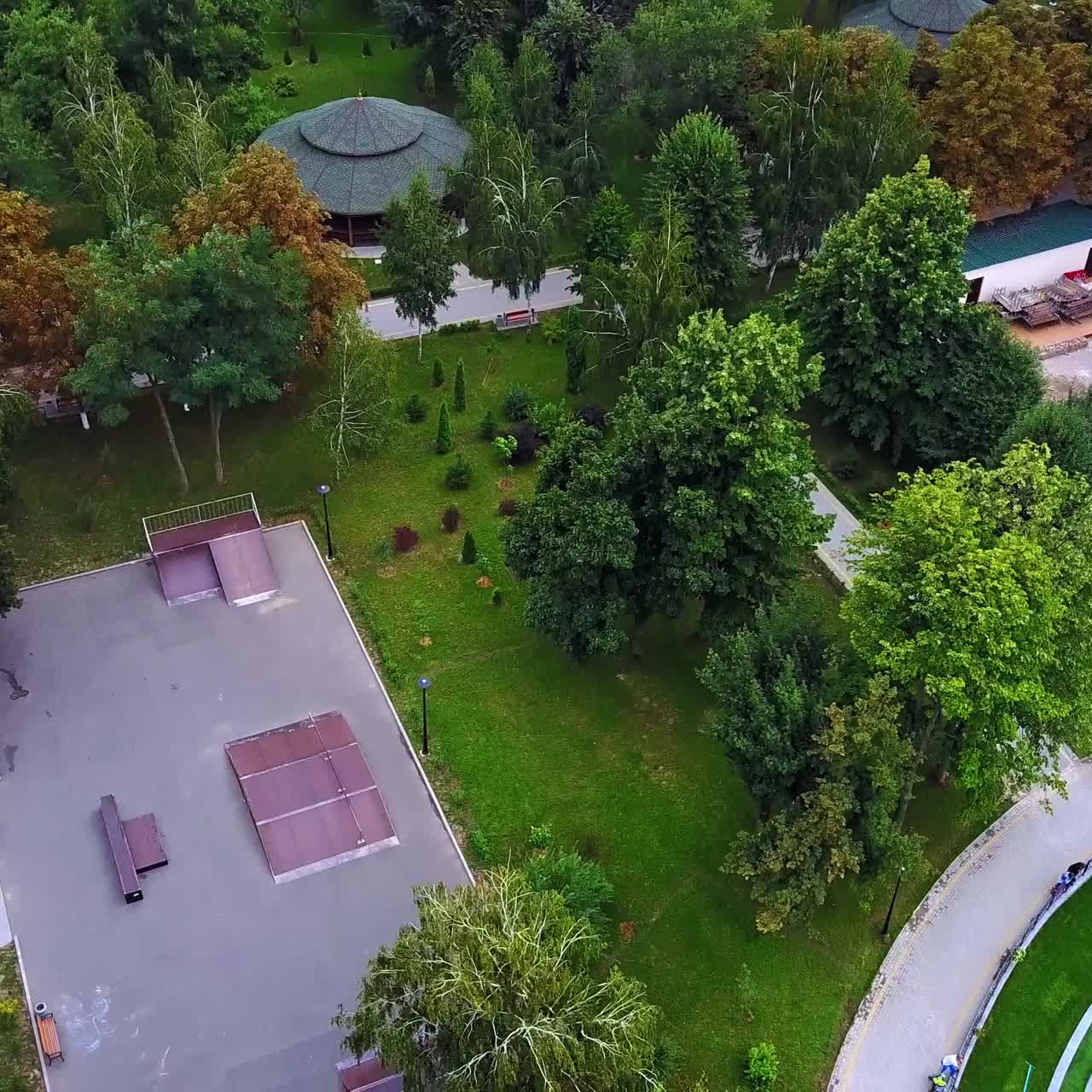 Neat territory of the city park with paths and pavilions. People standing around the oval pond with fountains. Aerial view