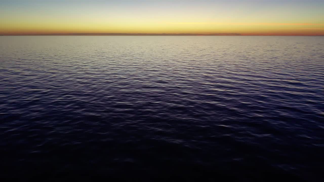 Calm ocean waters stretch to the horizon with subtle waves and a warm gradient sky transitioning from light to darker hues at sunset