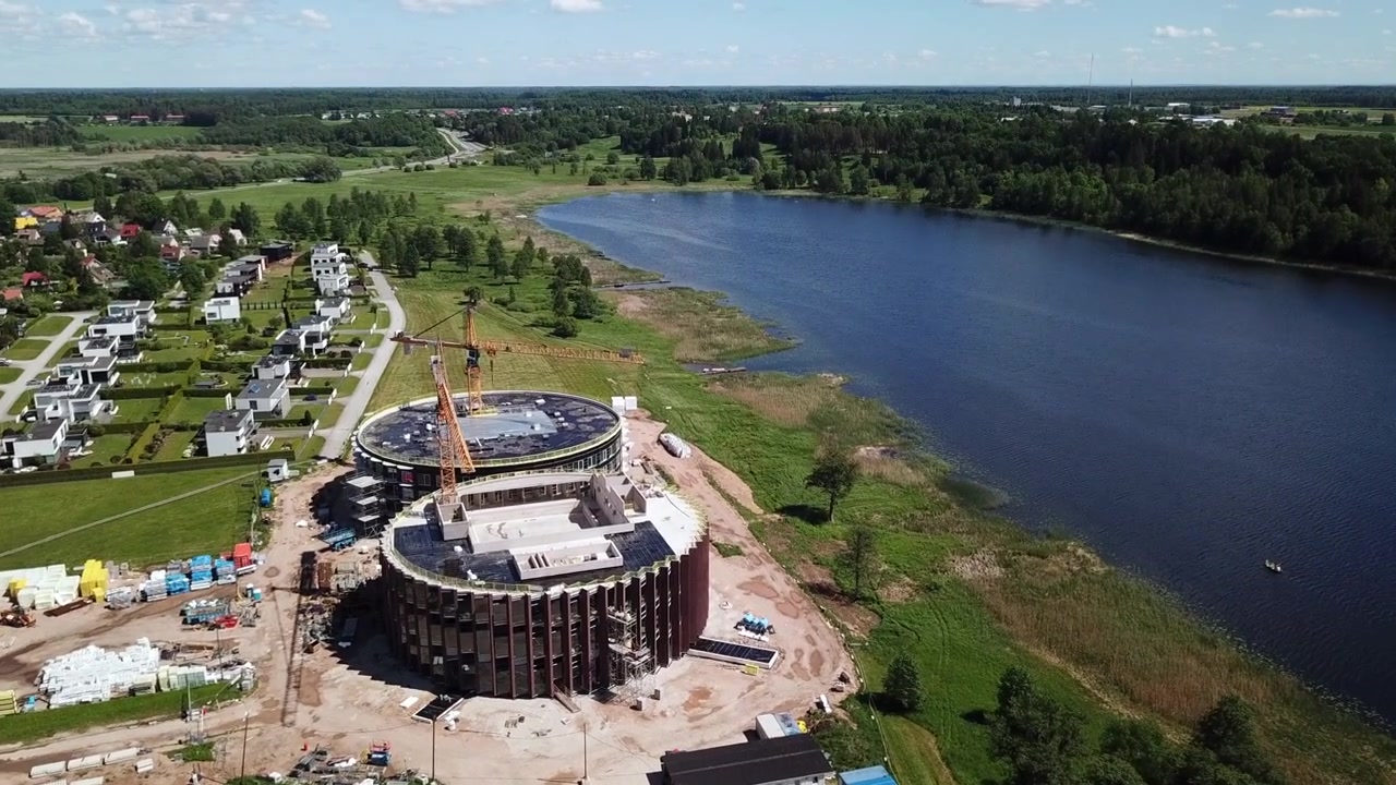 Panorama aéreo dinâmico orbitando o canteiro de obras do novo centro de spa moderno, com maquinário pesado e guindastes industriais ao lado do lago Viljandi