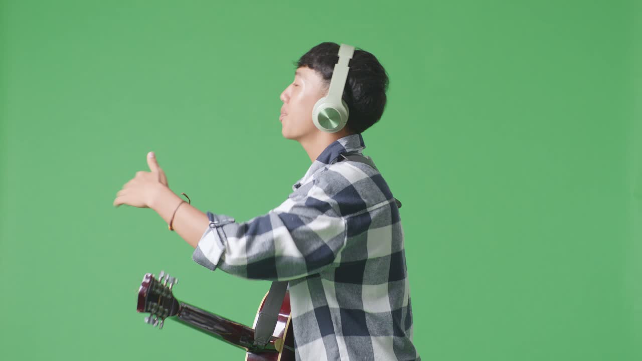 젊은 아시아 십대 소년이 기타와 함께 헤드폰을 착용하고 녹색 화면 배경에서 을 연주하는 측면 시각