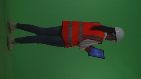 back view of full body asian female engineer with safety helmet looking at the wind turbine blueprint on a tablet while standing in the green screen background studio, during sunset or sunrise time