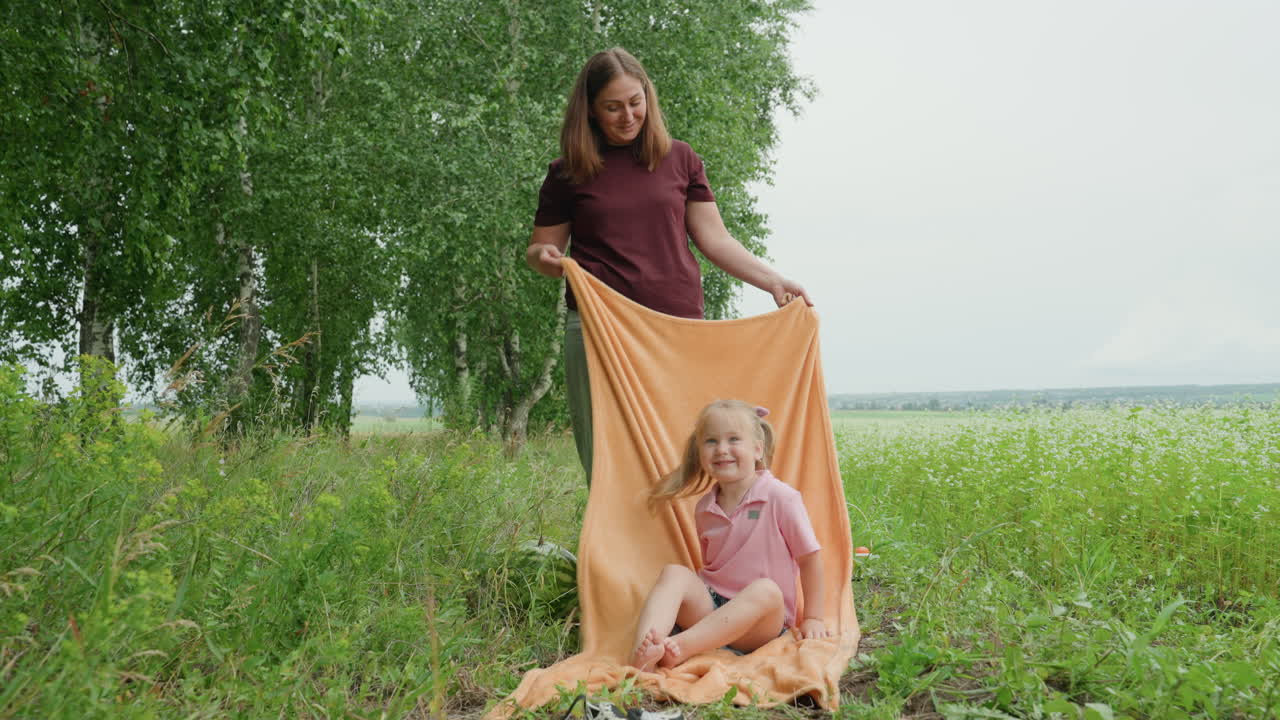 Weiße Mutter und Tochter im Freien, die eine Decke halten, Seeuferwiese mit Birkenkulisse, Kind sitzt auf orangefarbener Decke, Mutter drapiert Stoff, verspielte Pose, weiches Sommerlicht, offene Familie
