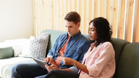 Young multiracial couple sitting on couch, using tablet and discussing documents together, at home