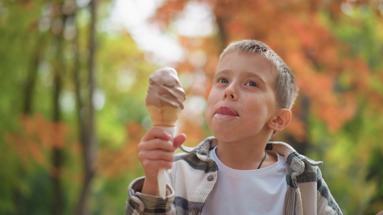 Niño disfrutando de un cono de helado al aire libre en un parque, sosteniendo el dulce y dando mordiscos, rodeado de follaje otoñal. El niño lleva una chaqueta a cuadros sobre una camisa blanca con iluminación suave.