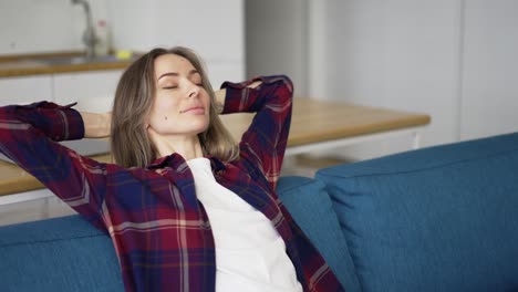 mujer joven tranquila relajándose en el sofá con los ojos cerrados las manos detrás de la cabeza