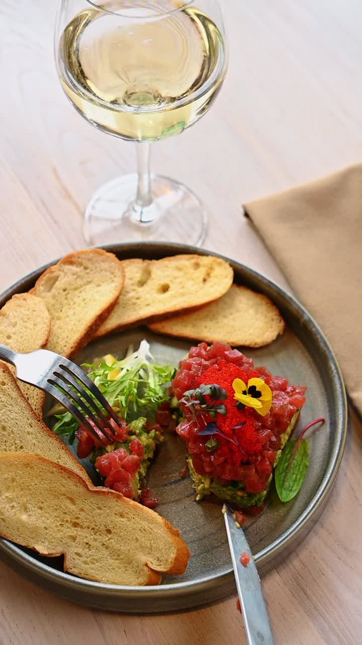 Persona che mangia tartare di tonno con forchetta e coltello. Primo piano di mani che usano forchetta e coltello per mangiare tartare di tonno con un bicchiere di vino bianco sullo sfondo.